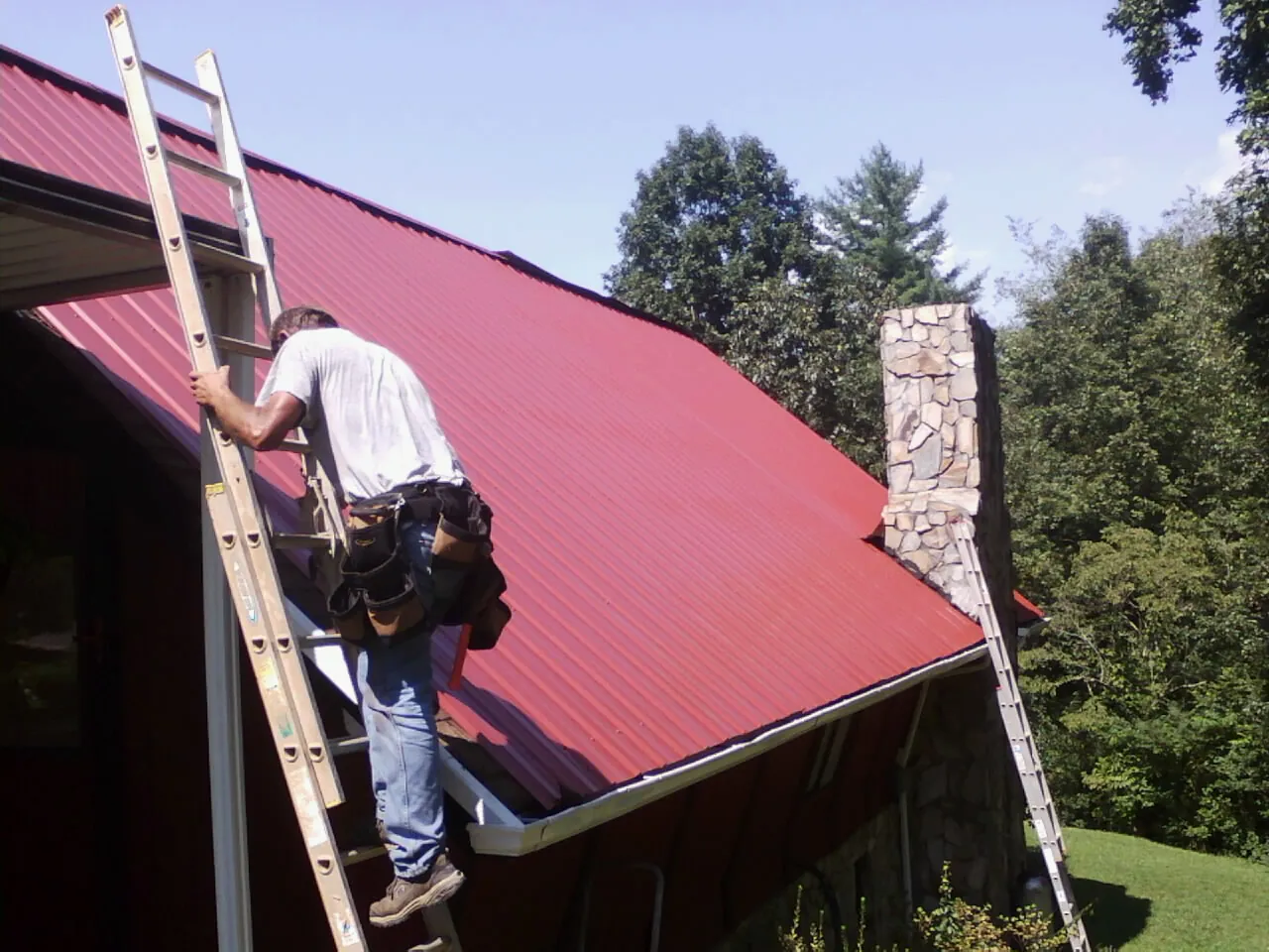 Roofer on a ladder beside a red metal roof and stone chimney — Soffit & Fascia Repair by Roofing Brothers in Roseville